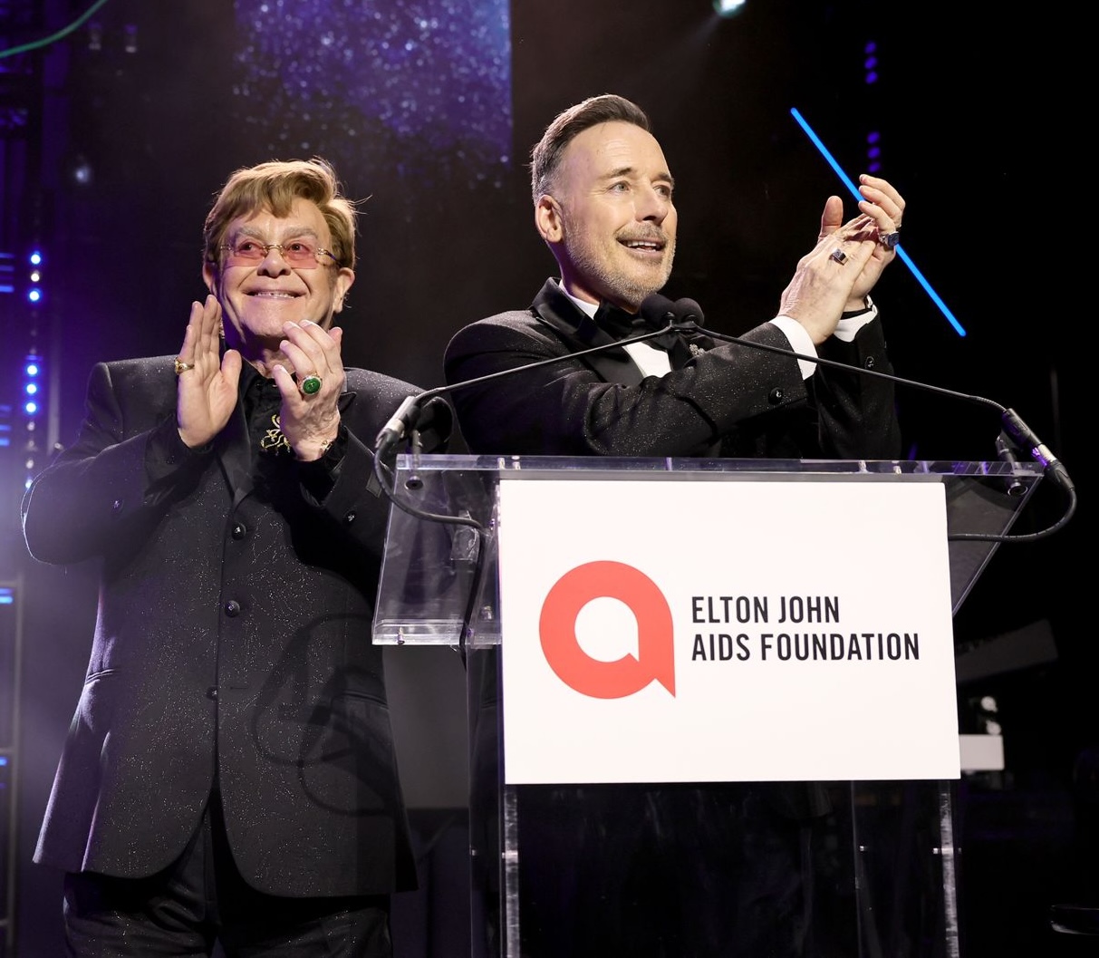 Elton John and David Furnish at podium with Elton John AIDS Foundation logo